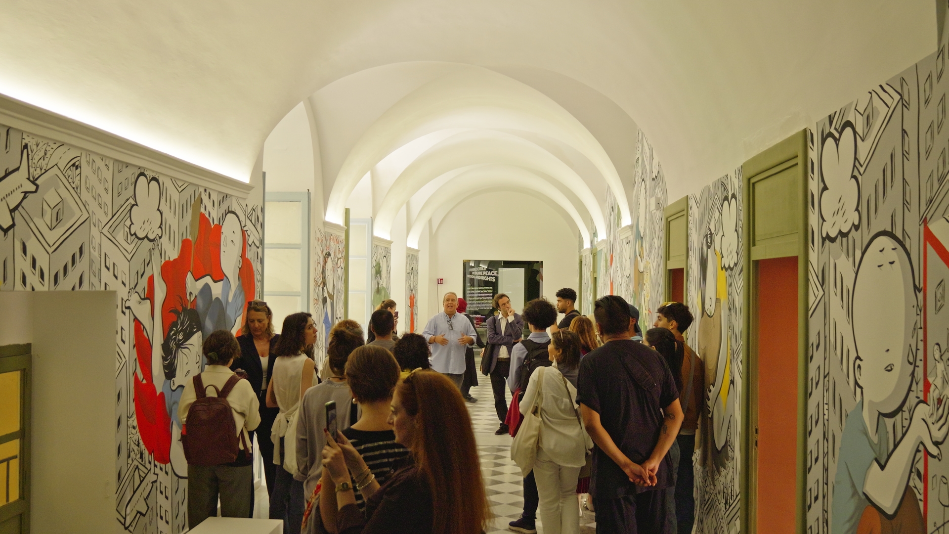 Un groupe de personnes écoute un homme dans un couloir orné de peintures murales.