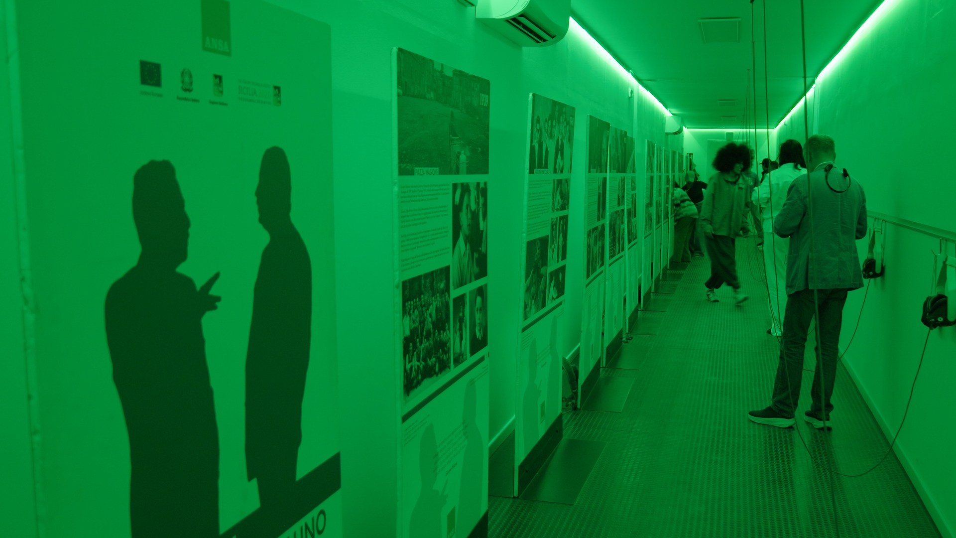 Sotto una luce verde, alcune persone partecipanti guardano affissi sulla storia della mafia.