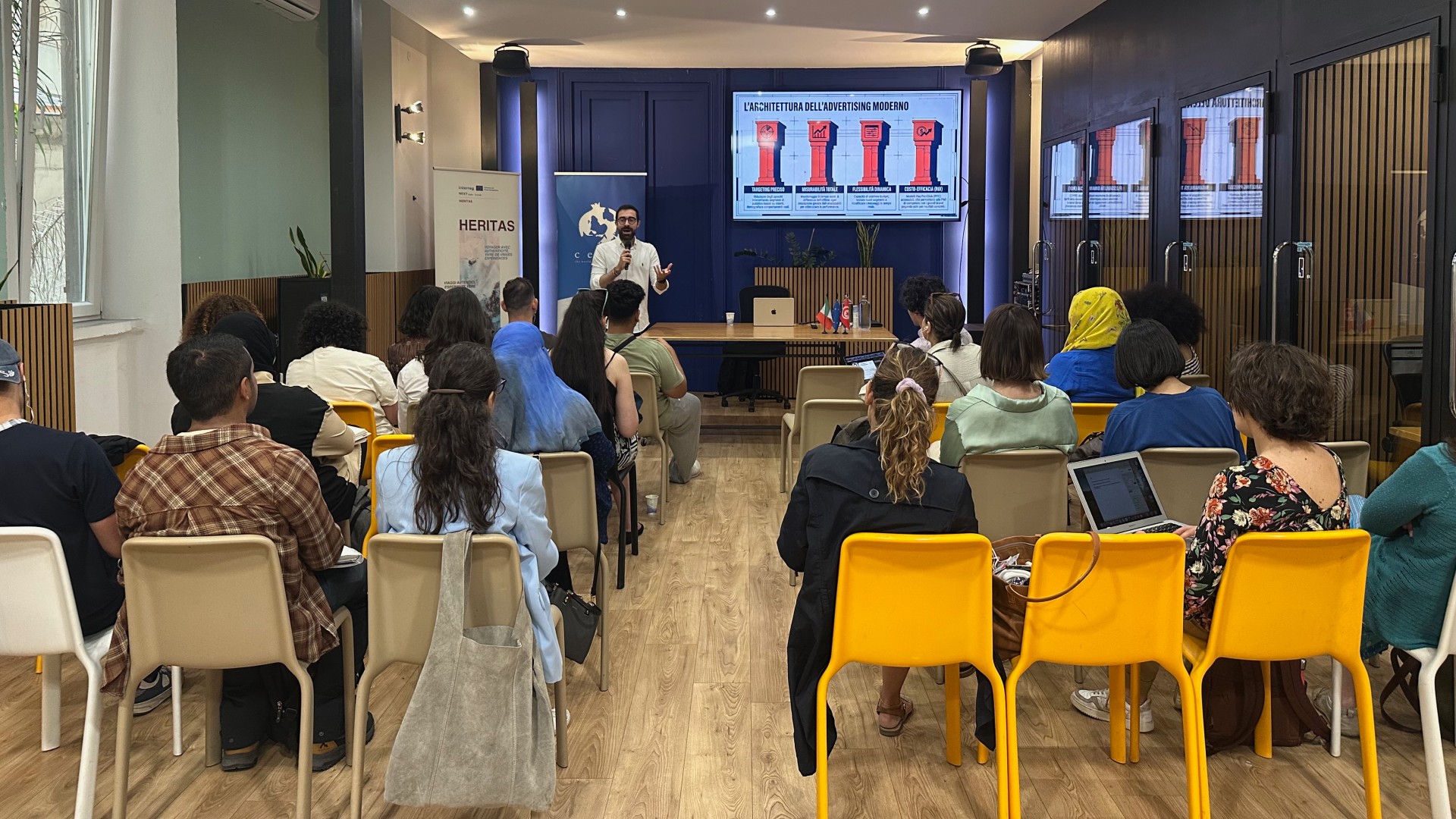 Un groupe de personnes écoute un homme présenter devant une présentation numérique.