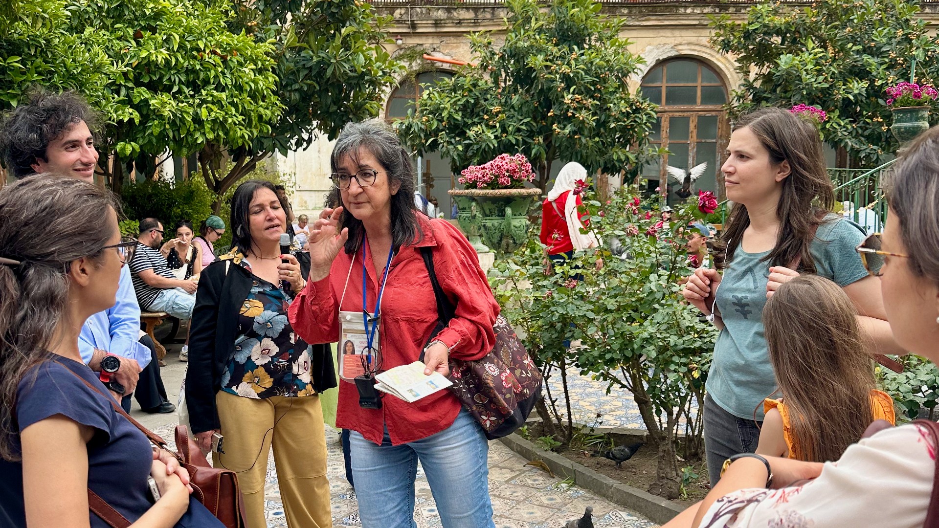 Une femme en chemise rouge s’adresse à un groupe de personnes au milieu d’un cloître.