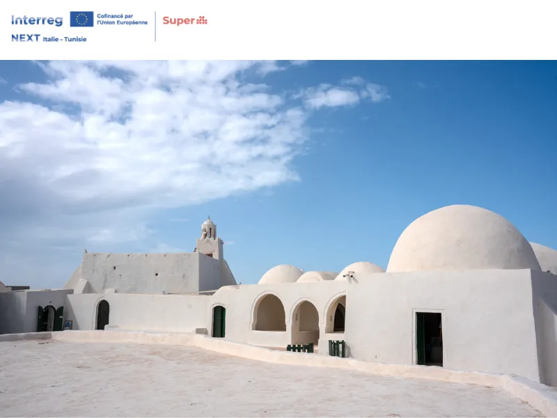 Veduta dell’architettura tradizionale di Djerba, in Tunisia, con edifici bianchi dalle cupole arrotondate e archi, sotto un cielo azzurro. L’immagine riporta i loghi del progetto SUPER – Interreg NEXT Italie-Tunisie.
